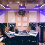后期混音师在双屏工作站前调整音频响度曲线，桌面上摆着监听音箱与笔记本，室内灯光柔和